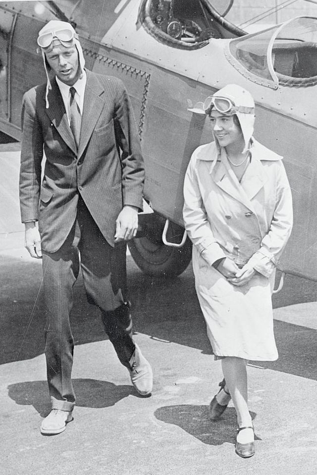 charles lindbergh and anne morrow lindbergh walk away from a two seat plane, both wear flying caps with goggles on their foreheads