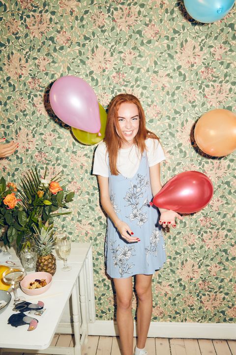 Portrait of cheerful young woman standing amidst balloons by table against wallpaper during party at home