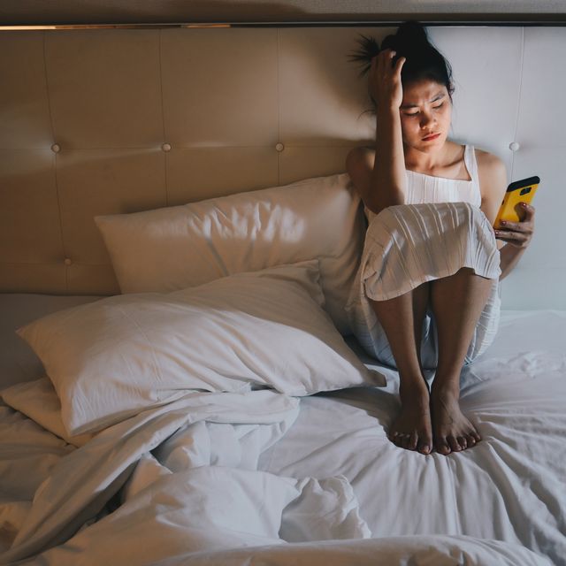 portrait of depressed woman sitting alone on the bed in the bedroom and looking to mobile phone in her hand