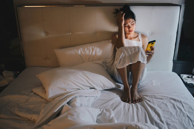 portrait of depressed woman sitting alone on the bed in the bedroom and looking to mobile phone in her hand