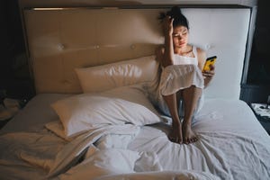 portrait of depressed woman sitting alone on the bed in the bedroom and looking to mobile phone in her hand