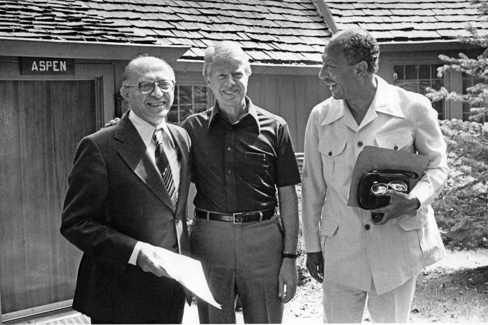 israeli prime minister menachem begin, us president jimmy carter, and egyptian president anwar al sadat stands outside a wooden building and smile, they wear business attire