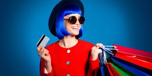 Portrait of glad woman in red outfit showing gesturing plastic credit card for comfortable quickly shopping in internet holding colorful packets in hand looking away isolated on blue background
