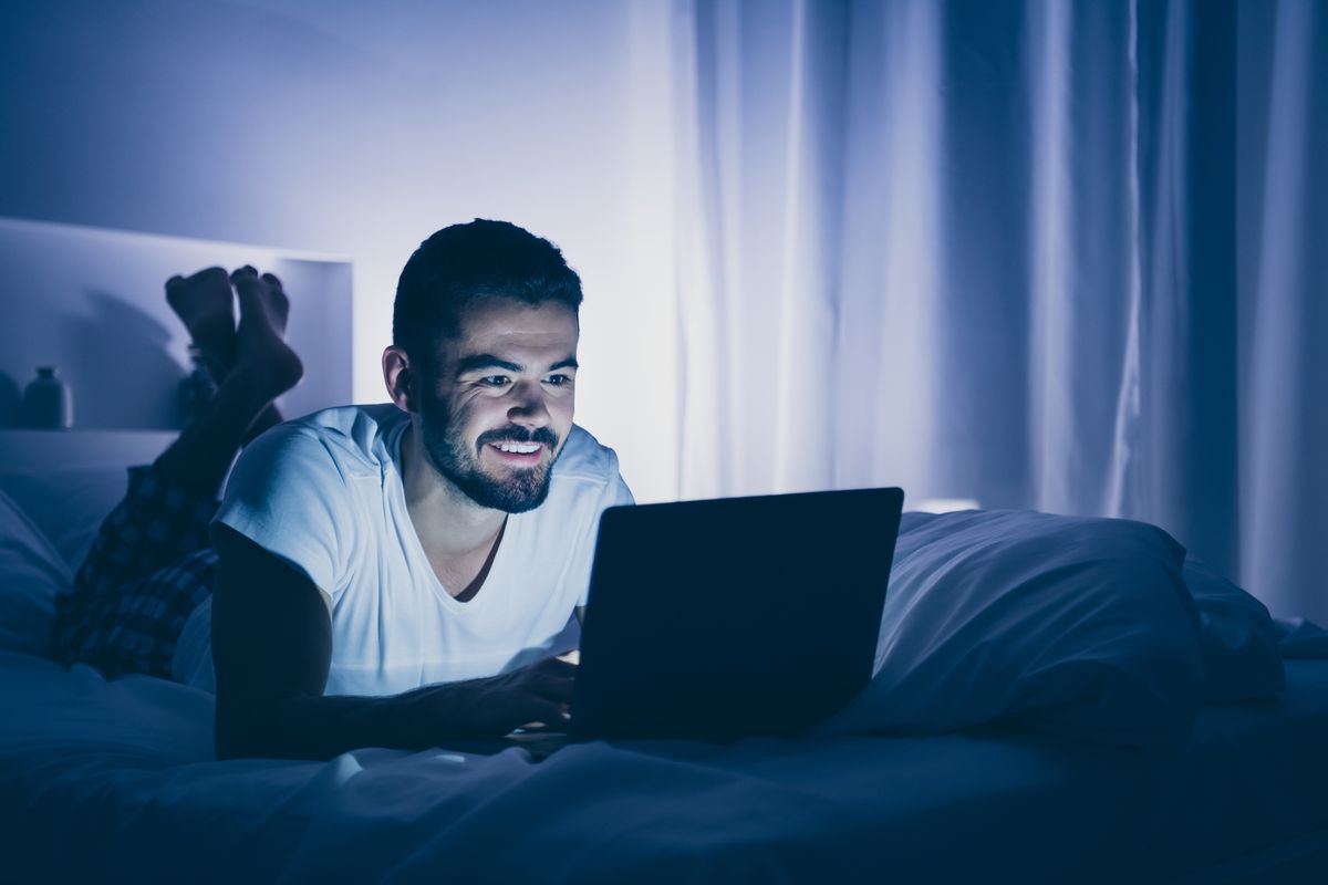 portrait of his he nice attractive cheerful cheery bearded guy lying on bed using digital laptop chatting spending time at night late evening home dark room flat house