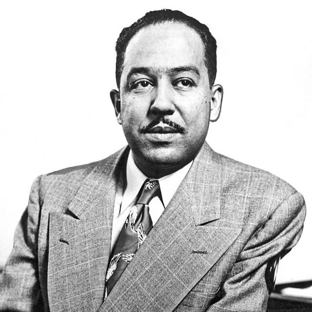 langston hughes sits and looks right of the camera, he wears a suit jacket, collared shirt, and patterned tie