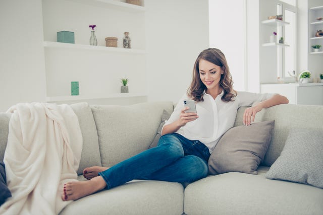 Portrait of pretty, charming, cheerful, trendy house wife in jeans, shirt with curly hair sitting on sofa having cell smart phone in hands chatting with lover searching contact using 3G wi-fi internet