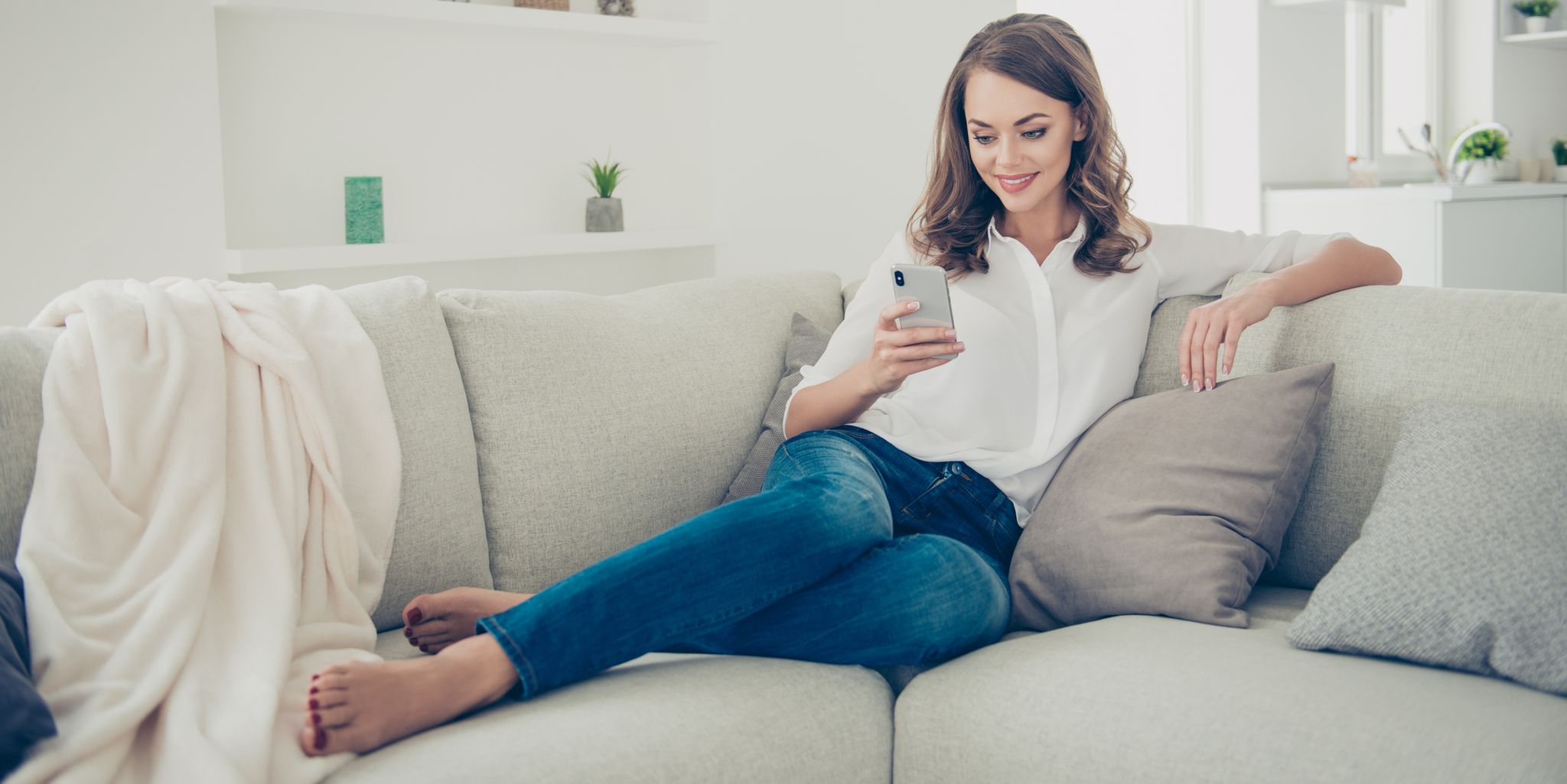 Portrait of pretty, charming, cheerful, trendy house wife in jeans, shirt with curly hair sitting on sofa having cell smart phone in hands chatting with lover searching contact using 3G wi-fi internet