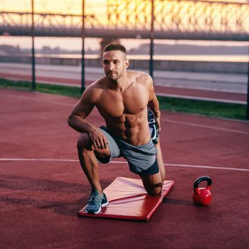 Portrait of shirtless muscular Caucasian man with serious face stretching leg while kneeling on the court. Next to him kettlebell.