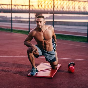 Portrait of shirtless muscular Caucasian man with serious face stretching leg while kneeling on the court. Next to him kettlebell.