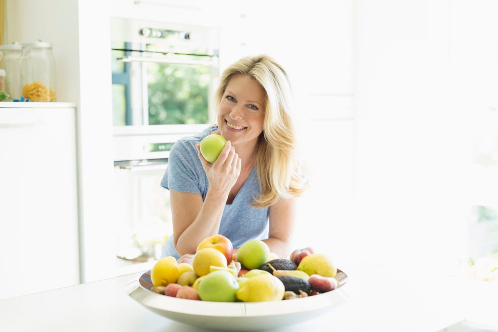 una chica rubia posa, con una manzana en la mano, frente a un frutero