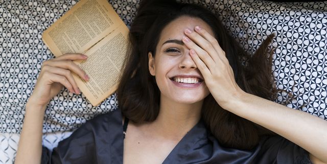Portrait of smiling young woman lying on bed with a book covering one eye with her hand