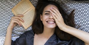 Portrait of smiling young woman lying on bed with a book covering one eye with her hand