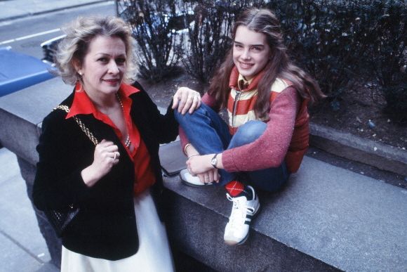 teri shields and brooke shields smile at the camera on a sidewalk, teri is wearing a red shirt underneath a black blazer and white bottoms, she clutches the strap of her purse that is hanging on her shoulder and rests her other hand on brookes knee, brooke is wearing an orange and yellow puffy vest, a red long sleeve shirt, jeans, argyle green and red socks and white sneakers, she is sitting on a concrete wall with her arms wrapped around her crossed legs