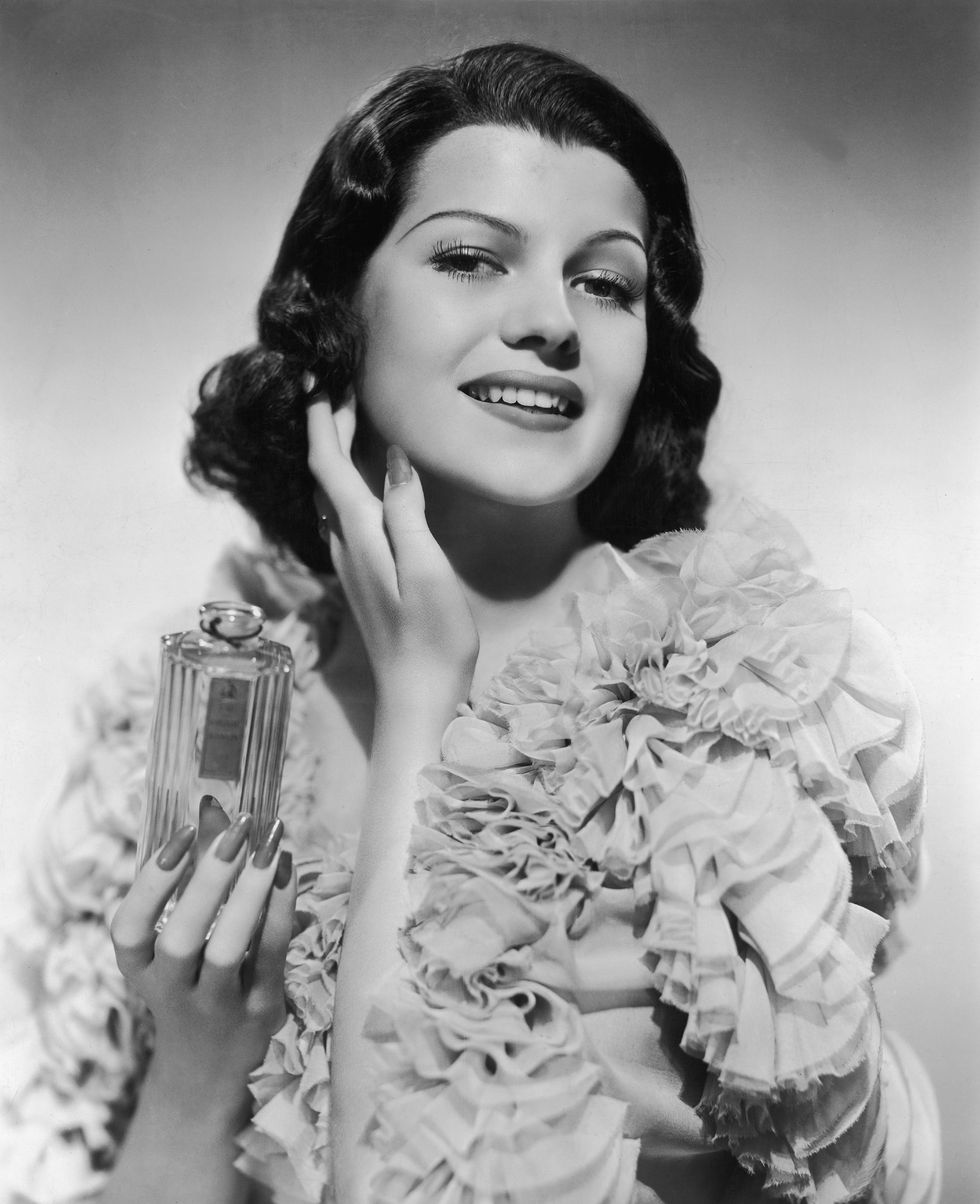 rita hayworth smiles at the camera while applying perfume to her neck, she wears a ruffled dress