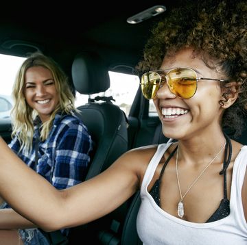 portrait of two women with long blond and brown curly hair sitting in car wearing sunglasses smiling