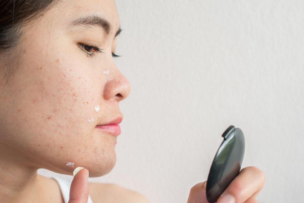 Portrait of young Asian woman applying acne cream/moisturizer on her face by looking a pocket mirror.