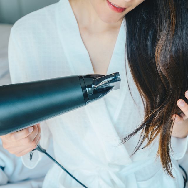 portrait of young asian woman using hair dryer for blows hot air to speed the evaporation of water to dry the hair in her bedroom