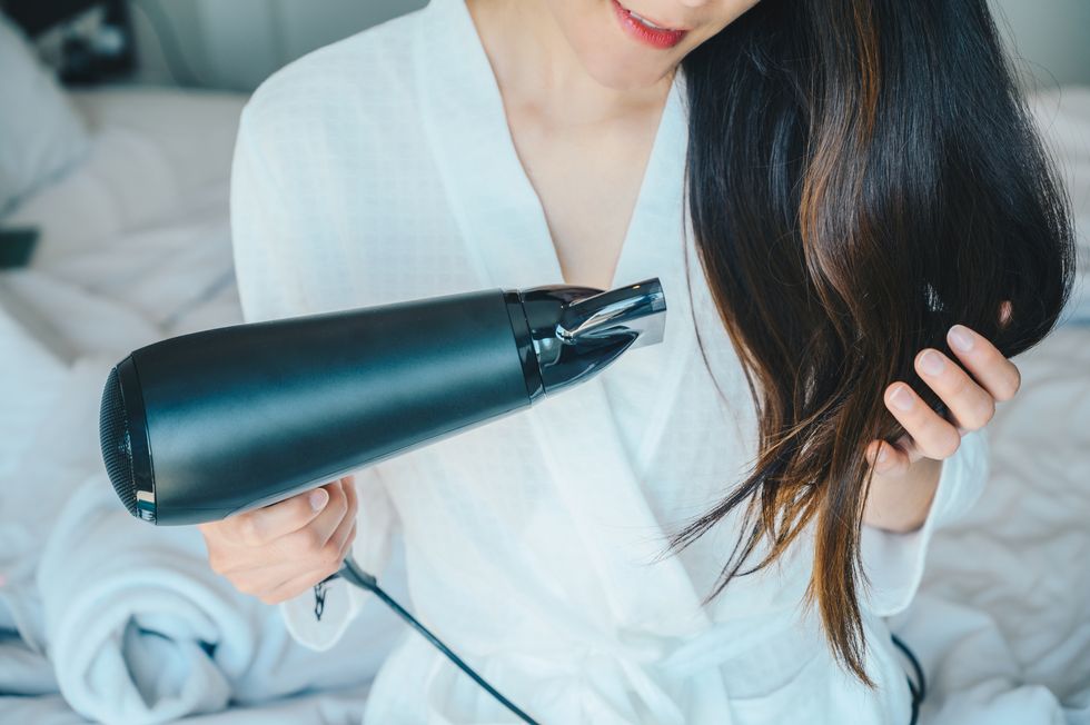 portrait of young asian woman using hair dryer for blows hot air to speed the evaporation of water to dry the hair in her bedroom