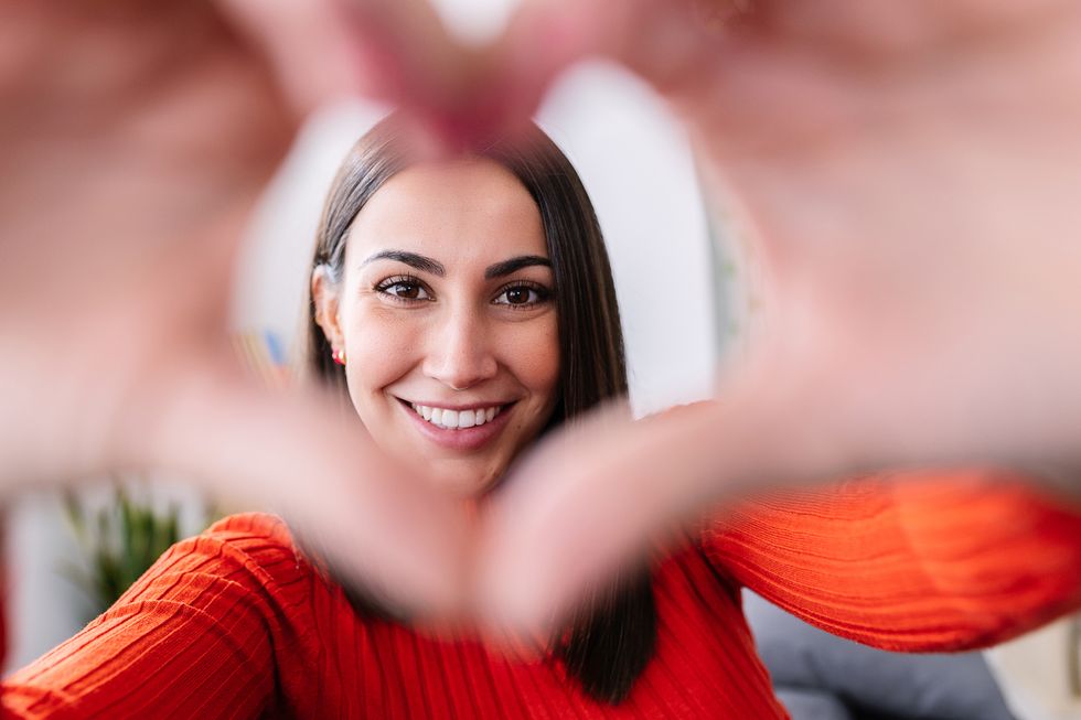portrait of young pretty woman showing shape heart with hands and looking through it with toothy smile while posing at her home love and happy people concept