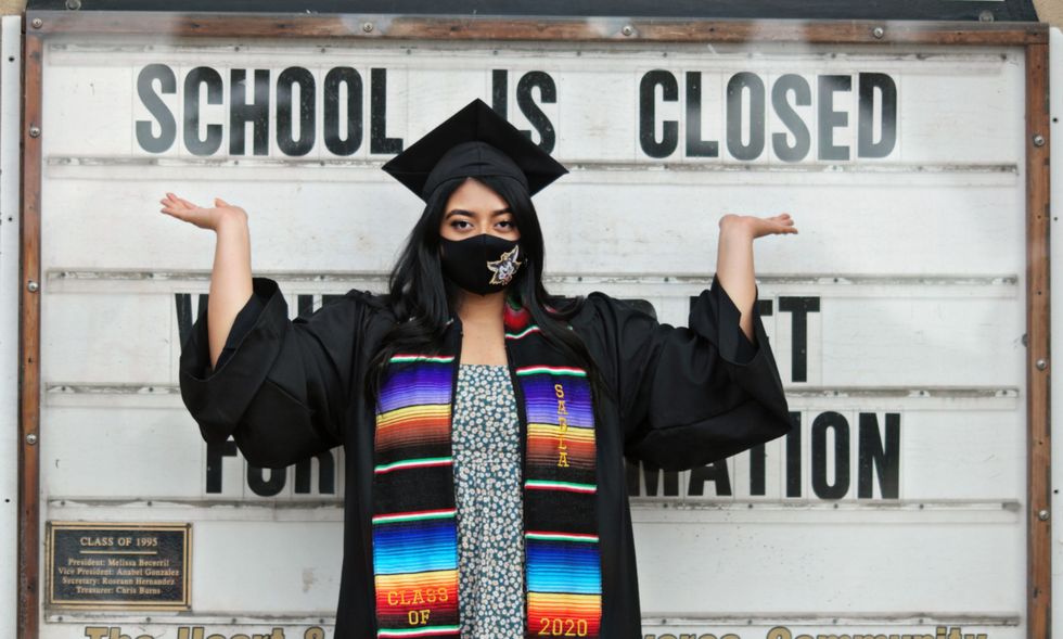 una ragazza in abito da laurea indica un muro con scritto "school is closed"
