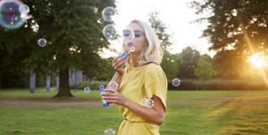 Portrait of young woman wearing yellow dress blowing bubbles in park at sunset