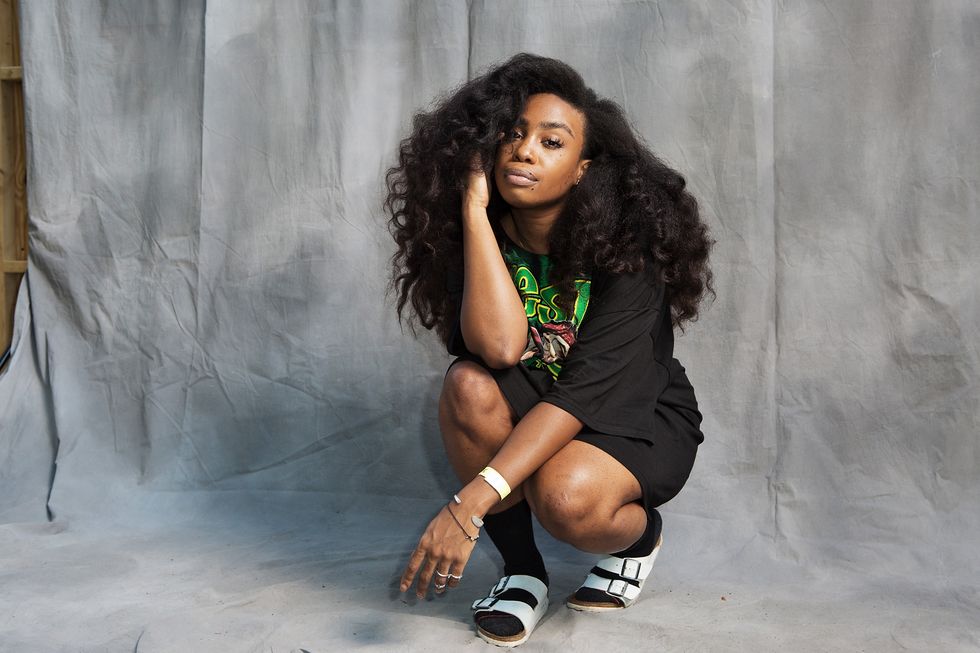 sza crouches and stares at the camera during a portrait session, she wears a black graphic tshirt, black socks, and white sandals