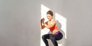 positive sportive woman with bun hairstyle and in tight sportswear doing squatting indoor studio shot illuminated by sunlight from window