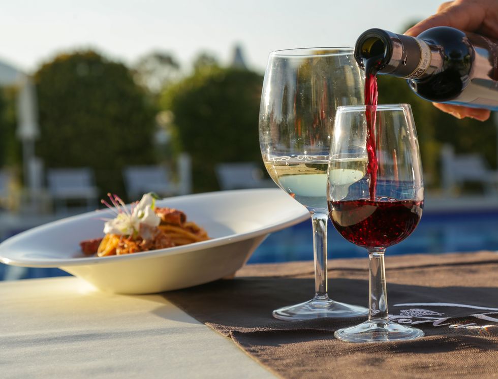 pouring a red wine from a bottle on a outdoor table with wine glasses and gourmet dish of pasta