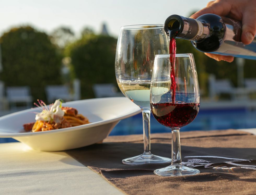pouring a red wine from a bottle on a outdoor table with wine glasses and gourmet dish of pasta