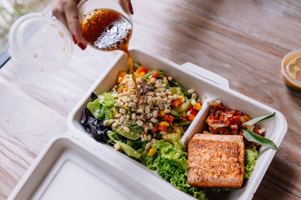 clean food meal box pouring salad dressing over grilled salmon fillet with tomato and bean sprout salad including carrot and corn