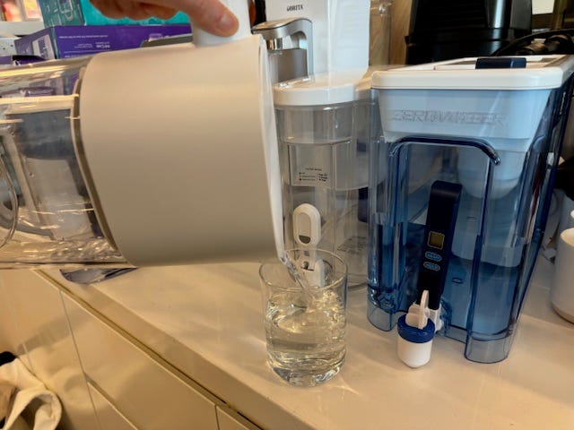 pouring water into a glass on a counter with other water filter pitchers in the background