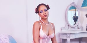 Rihanna wears a sheer pink teddy suit with over the knee sheer stockings, feathered heels, and purple eyeshadow. Her hair is done up in curlers. She sits on a lavender and gold lounge chair.
