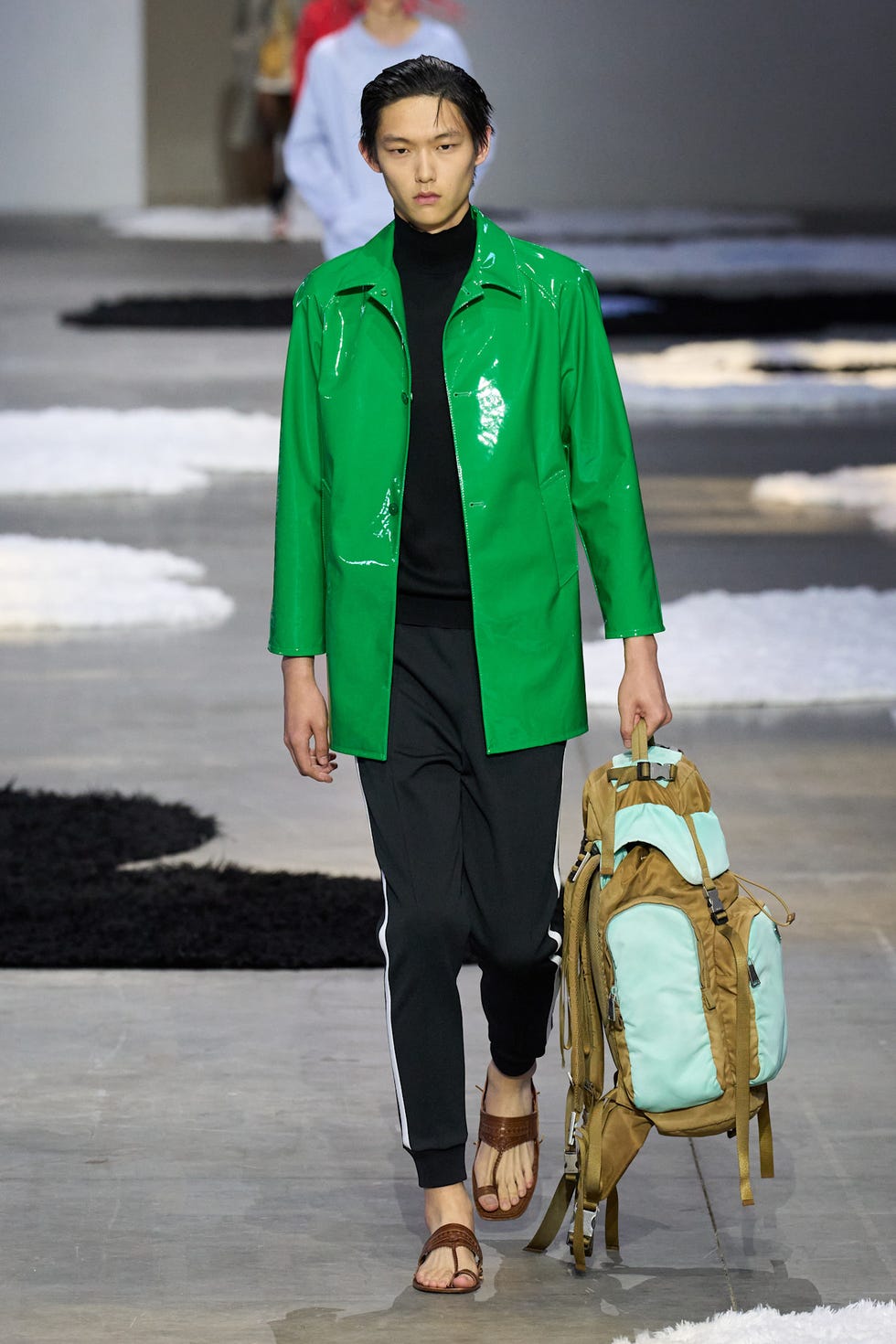 A model walks down the runway wearing a shiny green raincoat and holding a backpack