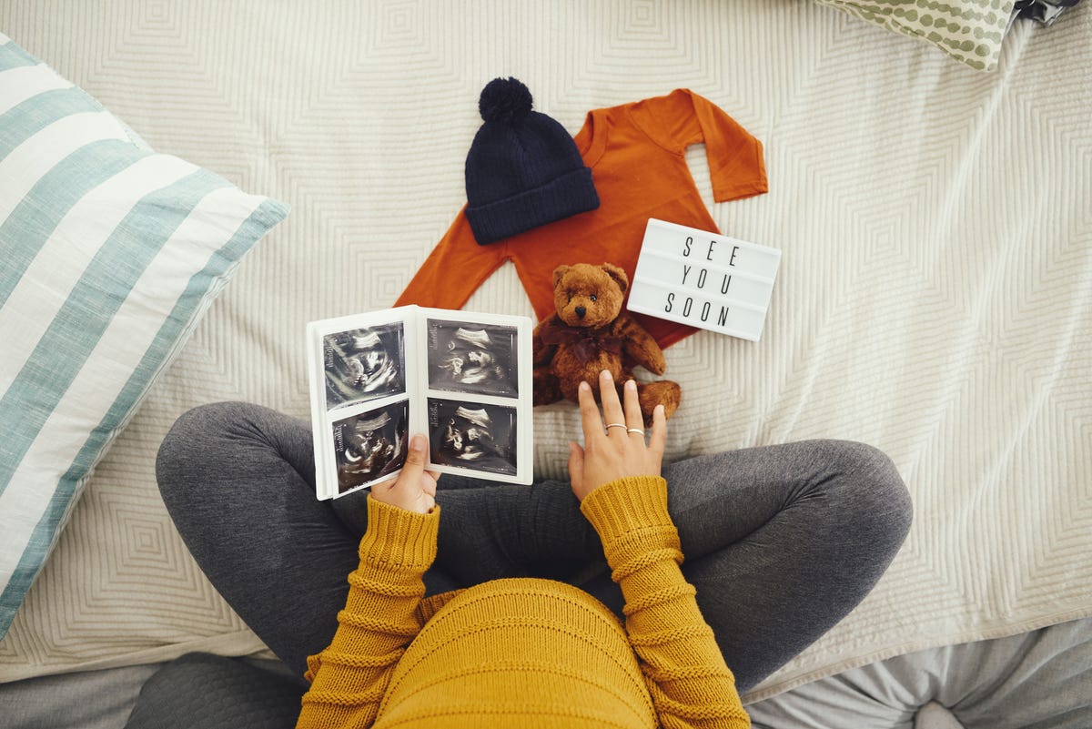 unrecognizable pregnant woman looking at a sonogram and baby clothing in her bedroom at home