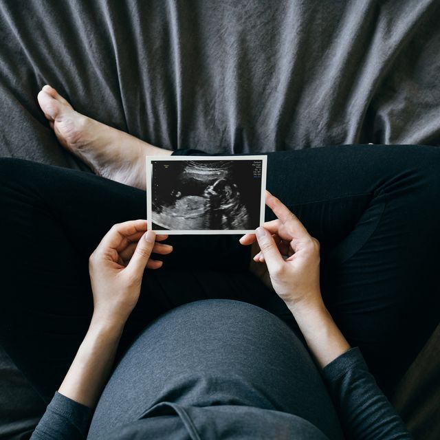 pregnant woman holding an ultrasound scan photo in front of her baby bump, sitting on bed at home