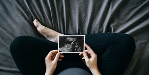 pregnant woman holding an ultrasound scan photo in front of her baby bump, sitting on bed at home