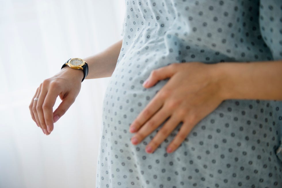 una mujer embarazada, con una bata de hospital y la mano en la tripa, controla con un reloj de muñeca sus contracciones