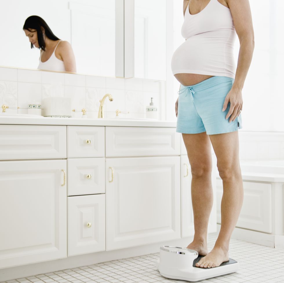 una chica embarazada, con pantalón corto y camiseta de tirantes, se pesa en unas báscula en su baño