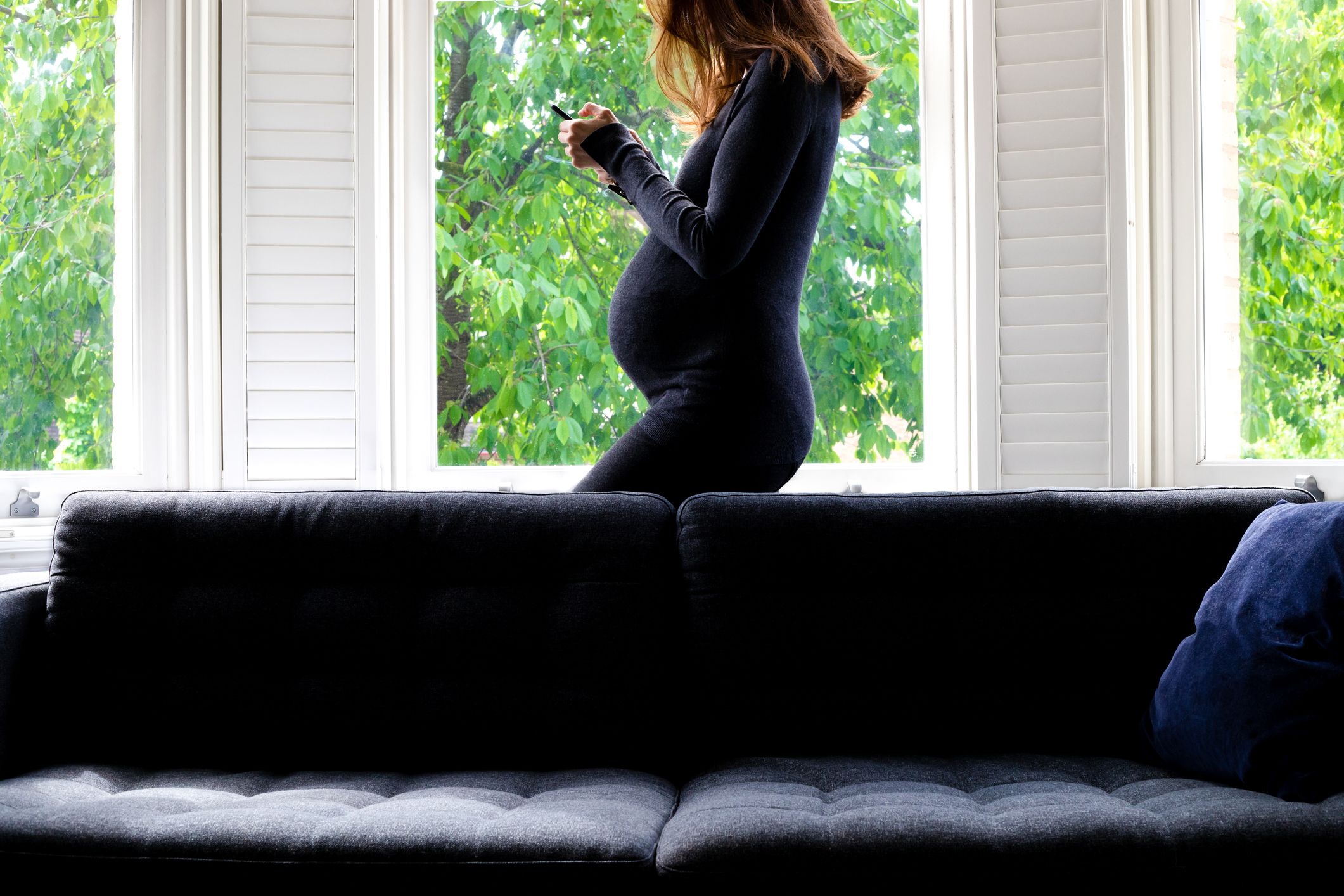 a pregnant woman using cell phone, standing by the window
