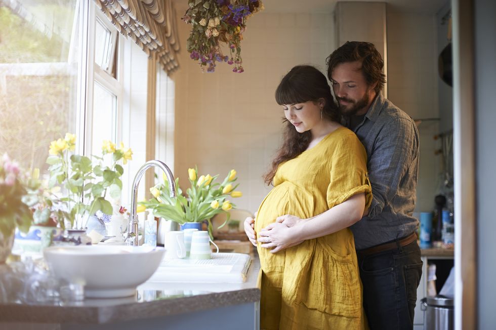 a pregnant couple embrace with their hands gently on stomach in the kitchen of their home with fresh spring flowers in vase