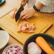 hands chopping raw chicken on a wooden cutting board with bowls of chopped vegetables nearby