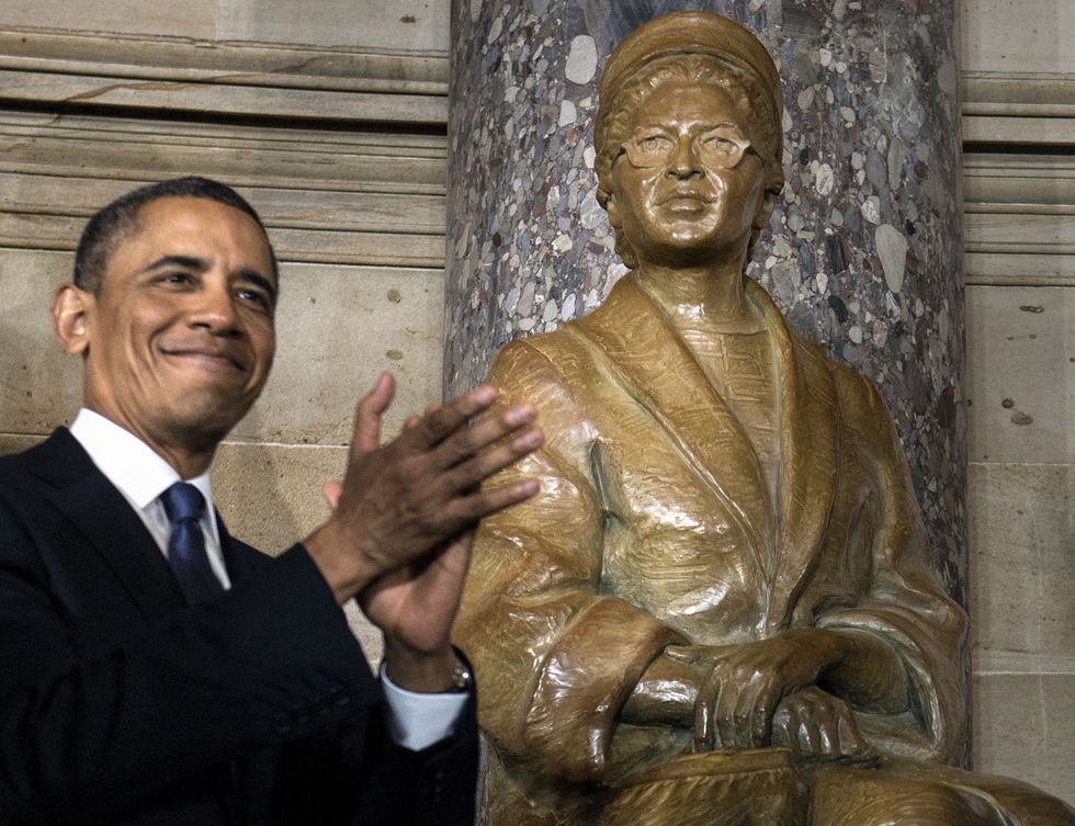 barack obama, wearing a black suit and blue tie, applauds and smiles while looking off camera, standing in front of a gold statue of rosa parks in front of a marble wall
