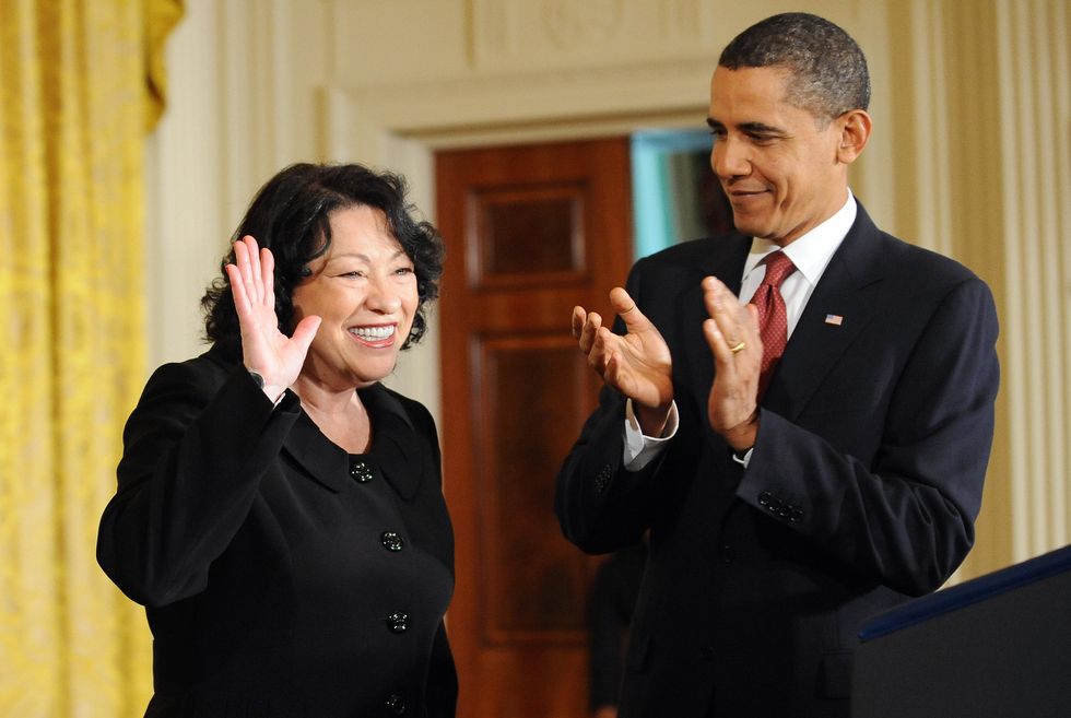 sonia sotomayor smiles and waves to an off camera crowd, earing a black dress, while barack obama smiles and applauds next to her, wearing a black suit and red tie