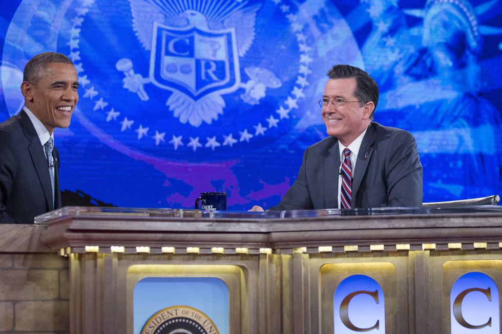 stephen colbert smiling while looking toward barack obama, also smiling, as they sit behind a table for an interview