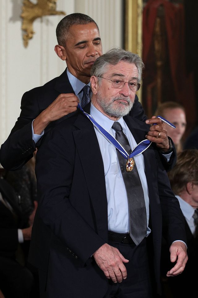 barack obama places a medal around robert de niros neck while both men stand inside a room, they both wear suits with blue collared shirts