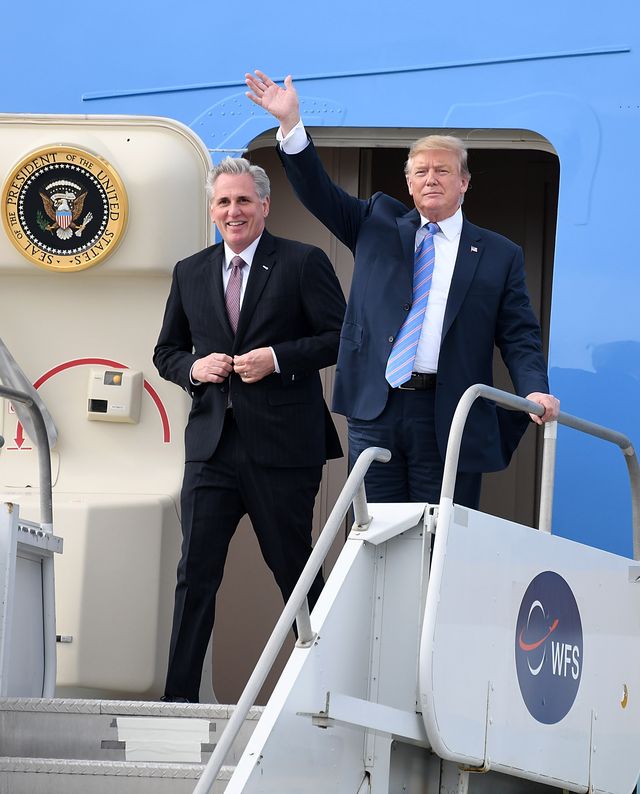 kevin mccarthy and donald trump walk out of an airplane door with a presidential seal on the inside of the door, mccarthy smiles and wears a suit and red tie, trump wears a navy suit with a blue tie and waves