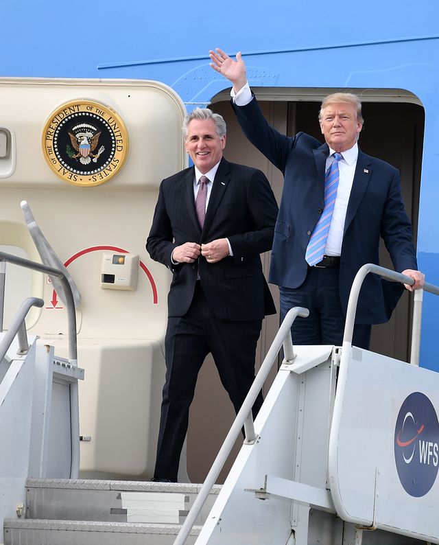 kevin mccarthy and donald trump walk out of an airplane door with a presidential seal on the inside of the door, mccarthy smiles and wears a suit and red tie, trump wears a navy suit with a blue tie and waves