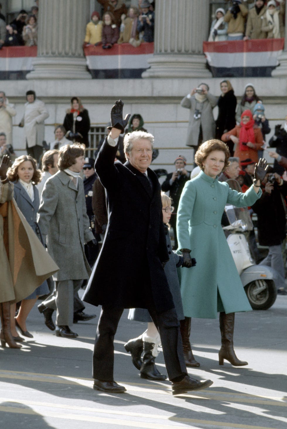 inaugural parade for president jimmy carter, 1977, jimmy walks on the street waving his right hand while he holds his daughter amy's hand in the left, rosalynn carter walks on amy's left side, holding one of the girl's hands and waving her left hand, in the background, other people are walking behind the carter family and still more people are watching as they walk by