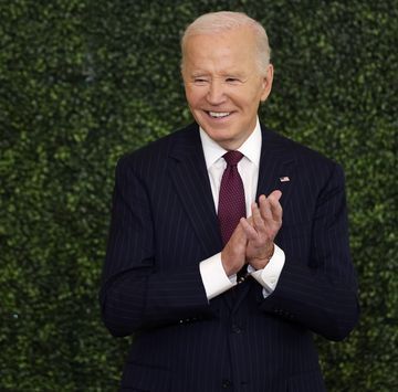 a man stands in a navy suit jacket white collared shirt and red tie, he claps his hands together in front of his chest and smiles while looking left