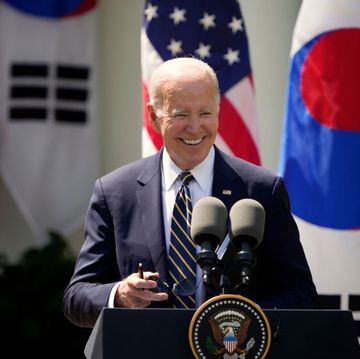 joe biden, wearing a dark blue suit jacket, white shirt, and striped tie, smiles while standing at a podium with two microphones on it, with american and south korean flags behind him